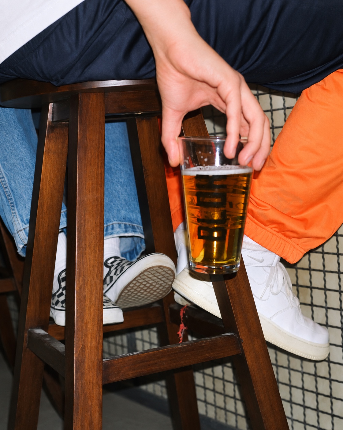 A hand holding a beer glass beside a wooden stool and white sneakers.